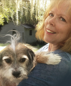 Smiling woman holding a small gray-and-white dog outdoors, with greenery and a shed in the background.