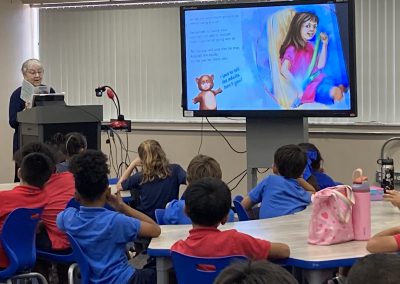 An older woman reads a children’s book to students seated at classroom tables while illustrated pages are displayed on a large screen.