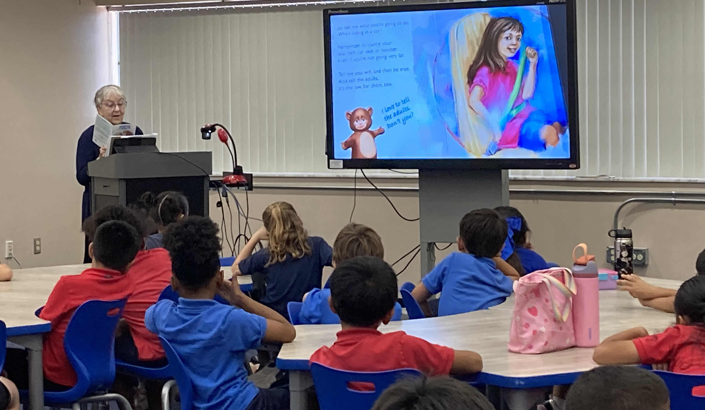 An older woman reads a children’s book to students seated at classroom tables while illustrated pages are displayed on a large screen.
