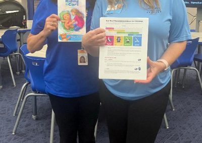 Two women stand in a classroom holding the book Riding in Cars and a child car-seat safety information sheet.