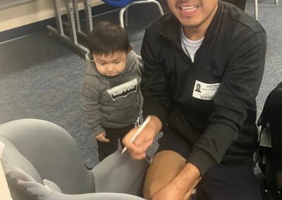 A man kneels beside a toddler and demonstrates a child car seat during a classroom safety program.