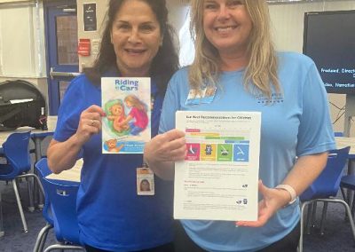 Two women stand in a classroom holding the book Riding in Cars and a child car-seat safety information sheet.