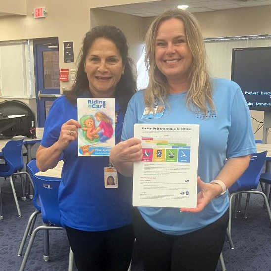 Two women stand in a classroom holding the book Riding in Cars and a child car-seat safety information sheet.