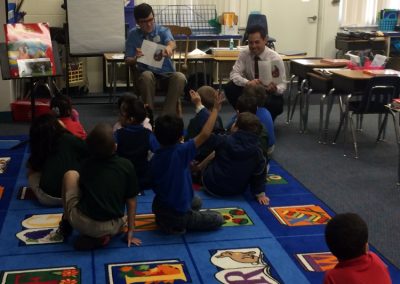Two men read a children’s book to a group of young students seated on a colorful classroom rug.