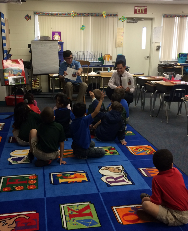 Two men read a children’s book to a group of young students seated on a colorful classroom rug.