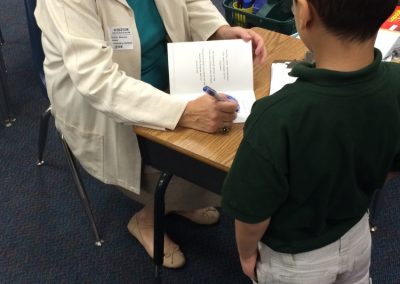 A woman signs a book for a young student at a classroom table.