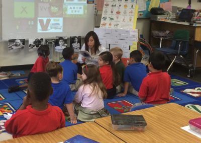 A woman reads to a group of elementary students seated on a classroom rug.
