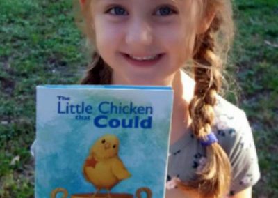 A smiling young girl holds a copy of The Little Chicken That Could outdoors.
