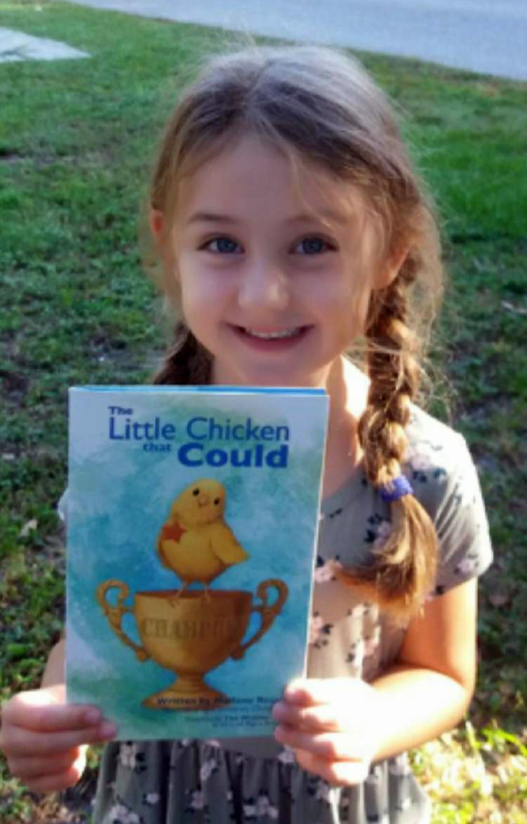 A smiling young girl holds a copy of The Little Chicken That Could outdoors.